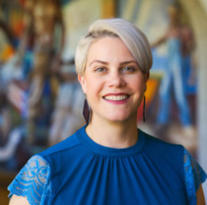 A woman with short blond hair and a blue top smiles before a mural background